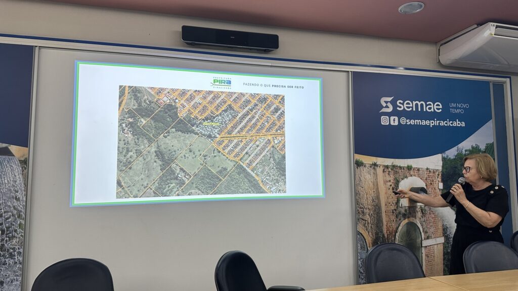 A imagem mostra uma mulher apresentando informações em uma sala de reuniões ou conferência. Ela está à direita da imagem, usando óculos e uma blusa preta, segurando um microfone com a mão esquerda e apontando para uma tela com a mão direita. Na tela, há um mapa aéreo, possivelmente de uma área urbana ou natural, com a palavra "PANTANAL" visível. Ao fundo, há uma parede branca com uma tela de projeção, acima da qual está escrito "PREFEITURA PIRA PIRACICABA FAZENDO O QUE PRECISA SER FEITO". Ao lado direito, um painel azul com o logotipo "semae UM NOVO TEMPO" e o perfil "@semaepiracicaba" indicam que o evento está ligado ao Serviço Municipal de Água e Esgoto de Piracicaba. O ambiente é iluminado por luz artificial e há uma mesa de madeira com cadeiras pretas na parte inferior da imagem. A apresentação parece estar relacionada a questões ambientais, urbanas ou de saneamento, considerando o mapa e as referências ao município.