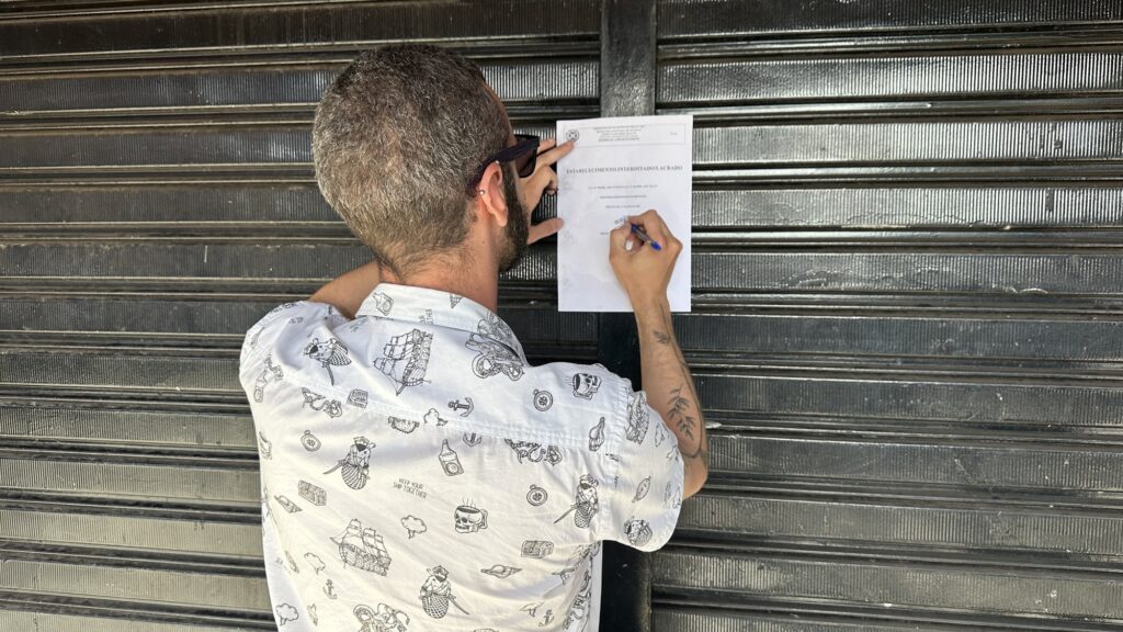 A imagem mostra um homem visto de costas, escrevendo em um documento oficial fixado em uma porta metálica escura, provavelmente um portão de enrolar. O documento tem o título "ESTABELECIMENTO INTERDITADO - LACRADO" e parece ser uma notificação administrativa em português. O homem tem cabelo curto e grisalho, usa óculos escuros presos na cabeça, um brinco na orelha esquerda e tem tatuagens no braço direito. Ele veste uma camiseta branca com estampas em preto, que incluem desenhos de navios, caveiras, sereias e a frase "KEEP YOUR SHIP TOGETHER". O ambiente sugere que o estabelecimento está fechado e interditado. A iluminação é natural e vem da frente, destacando o homem e o documento contra o fundo do portão metálico. A imagem transmite a ideia de uma ação oficial de fiscalização ou interdição em um local comercial.