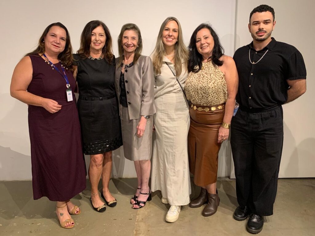 A imagem mostra seis pessoas alinhadas, posando para uma foto contra uma parede branca lisa. São três mulheres à esquerda, seguidas de uma mulher loira, outra mulher de cabelos castanhos e um homem à direita. A primeira mulher veste um vestido vinho escuro sem mangas e sandálias douradas, com um crachá pendurado no pescoço. A segunda mulher usa um vestido preto sem mangas com detalhes na barra e sapatos pretos baixos, também com crachá. A terceira mulher está com um terno cinza claro, com saia e blazer, e sapatos pretos de salto. A quarta mulher tem cabelos loiros e usa um vestido creme de mangas longas com tênis brancos. A quinta mulher veste uma blusa com estampa de oncinha, saia midi de couro marrom e botas marrons, com um cinto de corrente dourada. O homem à direita está vestido com uma camisa preta de mangas curtas, calça preta e sapatos pretos, usando um colar de pérolas e crachá. A iluminação é clara e uniforme, e o ambiente é simples, destacando as pessoas. A pose é formal, mas descontraída, sugerindo uma reunião social ou profissional.