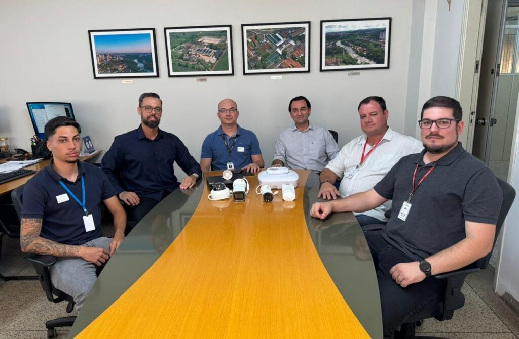 A imagem mostra seis homens sentados em torno de uma grande mesa oval em uma sala de reuniões ou escritório. Eles estão vestidos de forma casual, com camisas pólo e camisas sociais, e alguns usam crachás pendurados no pescoço. Sobre a mesa, há vários dispositivos eletrônicos, incluindo câmeras de segurança e um equipamento branco que parece ser uma base ou roteador. Ao fundo, na parede, há quadros com fotografias aéreas de áreas urbanas e industriais. A iluminação é clara e uniforme, e o ambiente parece profissional e organizado. Os homens parecem estar em uma reunião ou apresentação, com expressões neutras ou atentas. A mesa é de madeira clara e o ambiente tem cores neutras, como cinza e bege. À esquerda, é possível ver um monitor de computador e alguns materiais de escritório, indicando um espaço de trabalho funcional.