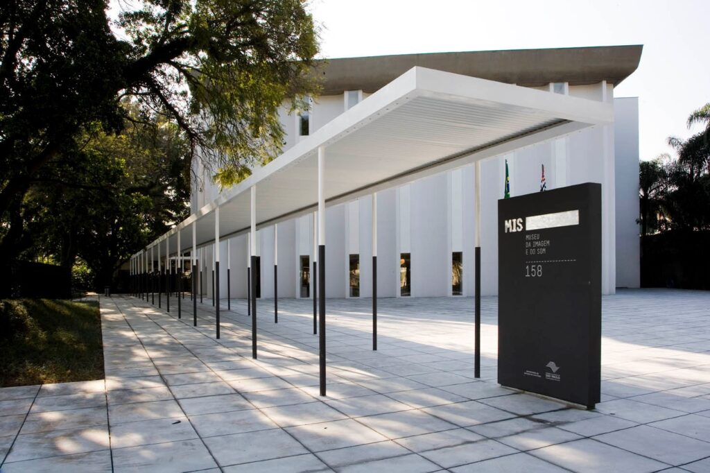 A imagem mostra a entrada do Museu da Imagem e do Som (MIS), localizado na cidade de São Paulo, Brasil. Em primeiro plano, há um monumento preto com inscrições em branco, onde se lê "MIS Museu da Imagem e do Som" e "Prefeitura do Município de São Paulo", além do número 158, provavelmente o endereço.À direita do monumento, há duas bandeiras, uma do Brasil e outra do estado de São Paulo, hasteadas. O prédio do museu é moderno, com paredes brancas e várias janelas estreitas e altas. Um longo toldo branco se estende da entrada do museu até o meio da imagem, sustentado por diversas colunas finas e escuras, criando um efeito de linhas paralelas que guiam o olhar em direção ao edifício.À esquerda, há árvores com folhagem verde que contrastam com o tom claro do piso e da estrutura do museu. O chão é revestido por grandes placas quadradas claras, formando um padrão geométrico. A iluminação é natural, com o sol projetando sombras das árvores e das colunas sobre o chão, indicando que a foto foi tirada durante o dia. A composição transmite uma sensação de modernidade, organização e tranquilidade.