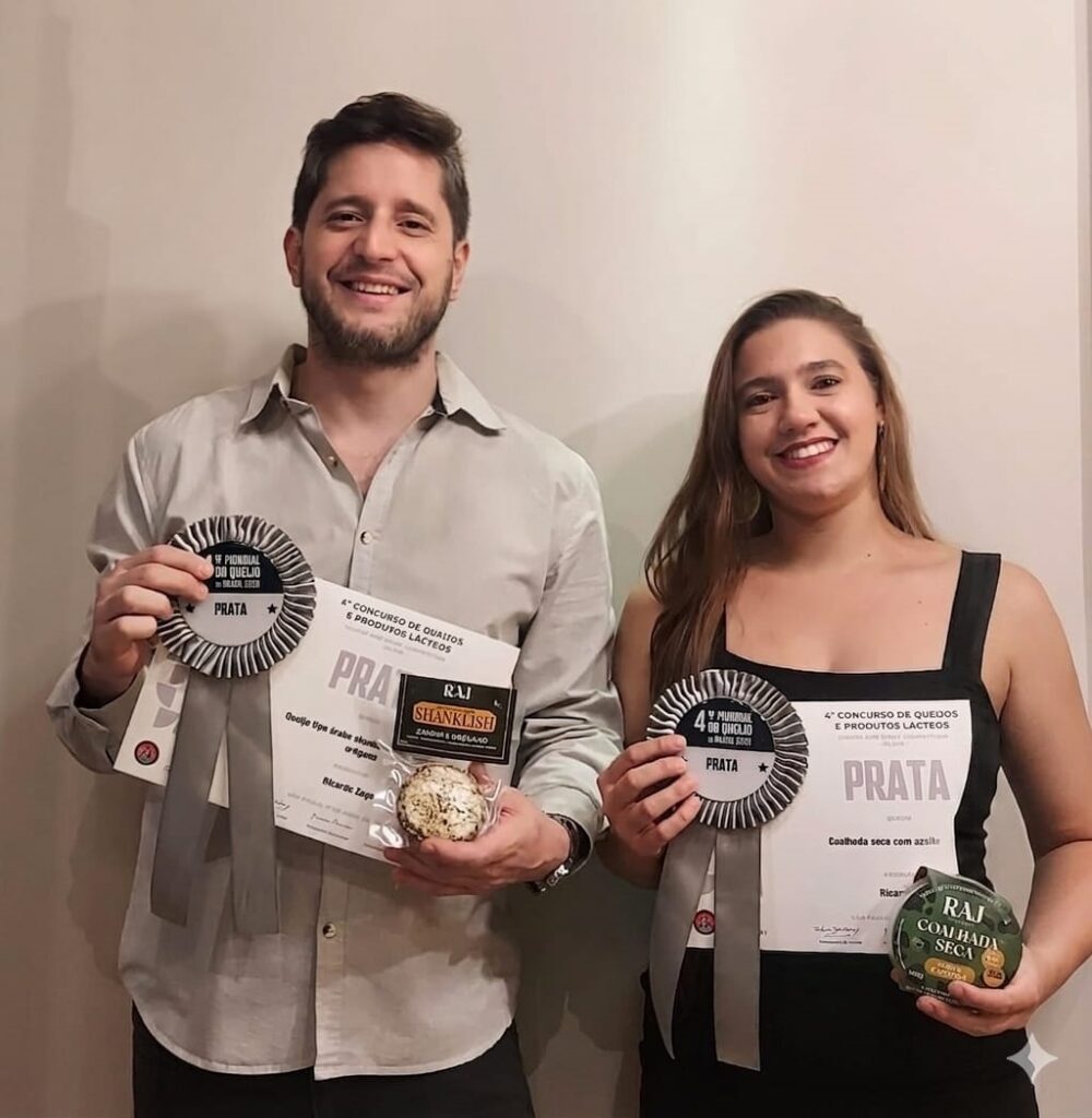 A imagem mostra duas pessoas, um homem e uma mulher, ambos sorrindo e segurando prêmios e produtos lácteos. Eles parecem estar em um evento ou cerimônia de premiação relacionada a queijos e produtos lácteos. O homem, à esquerda, veste uma camisa clara e segura em uma mão um certificado e um queijo redondo, e na outra mão uma fita de premiação de cor prata. A mulher, à direita, veste uma blusa preta sem mangas e também segura um certificado, um produto redondo (queijo) e uma fita de premiação prata. Os certificados indicam que eles receberam prêmios no "4º Concurso de Queijos e Produtos Lácteos", ambos na categoria "Prata". Os produtos que seguram têm rótulos com nomes que parecem ser "RAJ SHANKLISH" e "RAJ COALHADA SECA", que são tipos de queijos ou produtos lácteos. O fundo é simples e claro, destacando os premiados e seus produtos. A imagem transmite uma atmosfera de celebração e reconhecimento pela qualidade dos produtos apresentados.