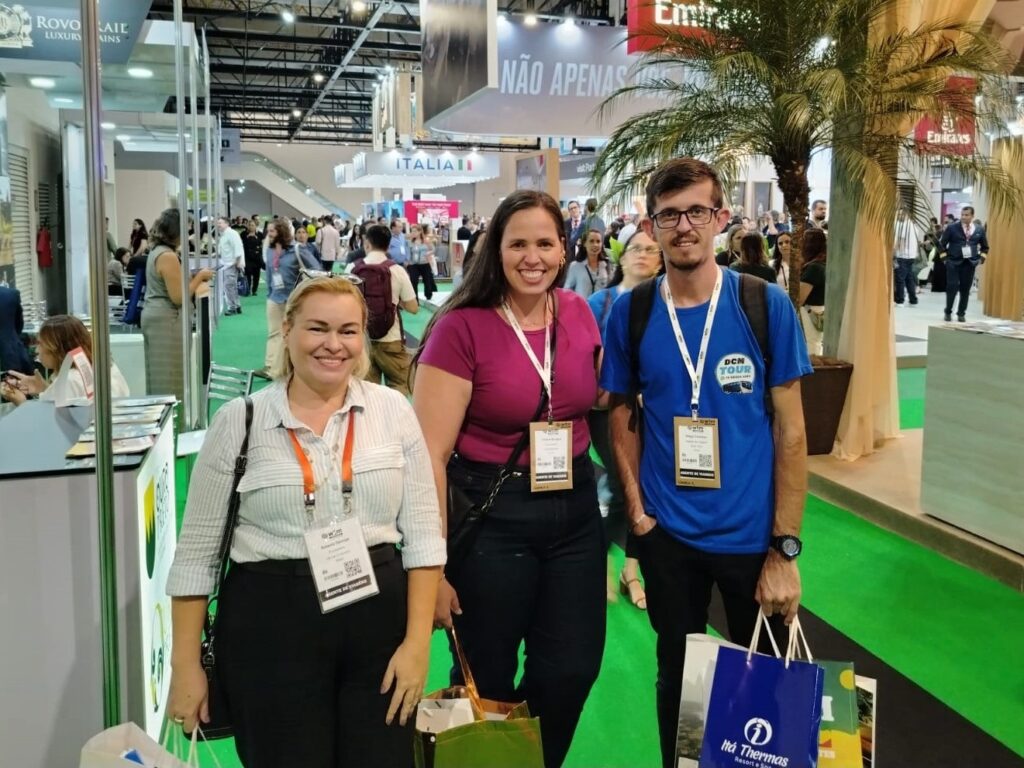A imagem mostra três pessoas posando para uma foto em um ambiente que parece ser uma feira ou exposição. Duas mulheres e um homem estão no centro da imagem, todos sorrindo e usando crachás pendurados no pescoço, indicando que são participantes ou expositores do evento. A mulher à esquerda veste uma blusa listrada e calça preta, a mulher no centro usa uma blusa rosa e calça preta, e o homem à direita está com uma camiseta azul, calça preta e mochila nas costas. Ao fundo, é possível ver outras pessoas circulando, estandes e algumas placas com nomes, como "ITALIAII", "Emirates" e "ROVO RAIL LUXURY". O chão é coberto por um carpete verde, e há uma planta (parece uma palmeira) à direita do grupo. A iluminação é clara e típica de ambientes internos de eventos, dando um ar profissional e organizado à cena. A foto tem boa qualidade e está focada nas três pessoas, com o fundo desfocado, mas ainda perceptível.