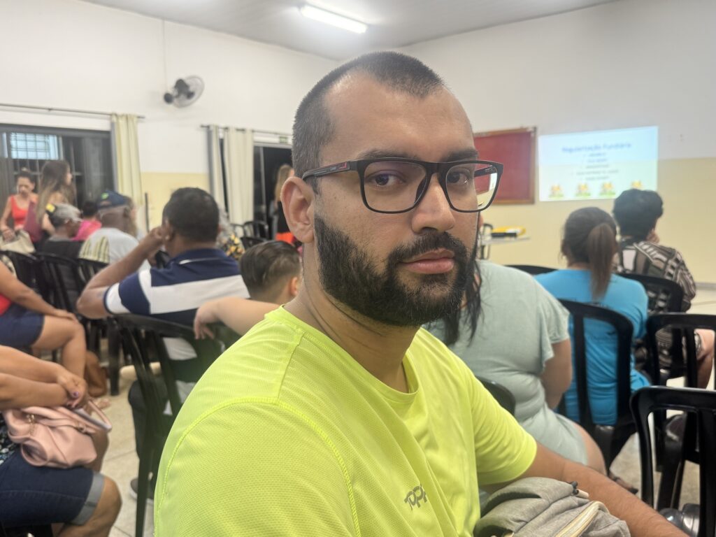 A imagem mostra um homem jovem, aparentando estar na faixa dos 20 a 30 anos, com cabelo curto e barba cheia escura. Ele usa óculos de armação preta e uma camiseta esportiva de cor verde neon com o logo "TOP" no peito. O homem está olhando diretamente para a câmera com uma expressão neutra. Ao fundo, é possível ver um ambiente que parece ser uma sala de reuniões ou aula, com várias pessoas sentadas em cadeiras pretas, de costas para a câmera. Na parede ao fundo, há um projetor exibindo um slide com o título "Regularização Fundiária", sugerindo que o local está sendo usado para uma apresentação ou palestra sobre esse tema. A sala tem paredes brancas, piso de cerâmica clara e um ventilador de parede, com iluminação clara e uniforme. A imagem transmite um contexto de evento comunitário ou educacional.