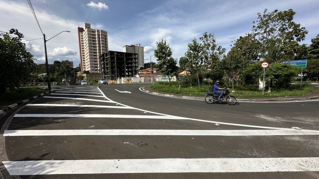 A imagem mostra uma cena urbana durante o dia, com um amplo cruzamento para pedestres em primeiro plano. O chão é asfaltado e o faixa de pedestres é pintada em branco, destacando-se contra o asfalto escuro. Um motociclista, vestido com camiseta azul e capacete, está atravessando a rua. Ao fundo, há diversos prédios, incluindo um edifício moderno de cor avermelhada e outro em construção ou parcialmente inacabado. Árvores verdes e um céu azul com poucas nuvens completam o cenário, trazendo um contraste natural ao ambiente urbano. À direita da imagem, há uma placa de trânsito circular vermelha indicando uma seta para a direita, e uma placa verde com os dizeres "Av. Independência" e "São Paulo", indicando que a cena se passa em uma avenida dessa cidade. No geral, a imagem transmite uma atmosfera típica de uma rua movimentada em uma cidade brasileira, com elementos urbanos e naturais bem equilibrados.