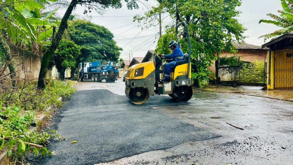 A imagem mostra uma cena de pavimentação de rua em uma área provavelmente residencial. No centro da imagem, há um compactador de solo amarelo e cinza da marca WACKER R SON, modelo D12, que está sendo operado por uma pessoa vestindo uniforme azul e capacete. O compactador está passando sobre uma camada recém aplicada de asfalto, que ainda parece fresca e úmida. À esquerda, há um caminhão azul com a cabine branca estacionado, que possivelmente está sendo usado para transportar o material de pavimentação. Ao fundo, é possível ver algumas casas com telhados de cerâmica e vegetação ao redor, incluindo bananeiras e outras plantas. O céu está nublado, sugerindo um dia com clima fechado, e o chão parece molhado, indicando que pode ter chovido recentemente. A imagem transmite uma sensação de trabalho em andamento, com destaque para o maquinário e o processo de pavimentação.