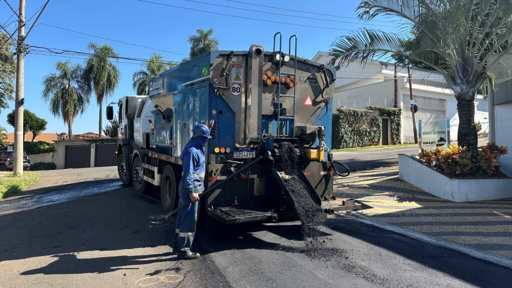 A imagem mostra uma cena de pavimentação de rua em um dia ensolarado. No centro, há um caminhão azul e prata especializado em aplicação de asfalto, com o asfalto quente sendo despejado na pista a partir da parte traseira do veículo. Um trabalhador vestido com uniforme azul e capacete está ao lado do caminhão, segurando uma pá, aparentemente supervisionando ou auxiliando no processo de pavimentação. Ao fundo, há árvores de palmeira e algumas construções, incluindo um prédio com uma placa que diz "SMART SENSING" e "SMART AGRI", indicando que o local pode estar relacionado à agricultura inteligente. O caminhão possui uma placa de licença com o número "UDS4A95" e sinais de alerta, como "PERIGO" e um símbolo indicando limite de velocidade de 80 km/h. A luz do sol cria sombras marcantes do caminhão e do trabalhador sobre o asfalto recém-aplicado, destacando a textura granulada do material. A cena transmite uma sensação de trabalho em andamento e cuidado na manutenção ou construção da via, em um ambiente que mistura elementos urbanos e naturais.