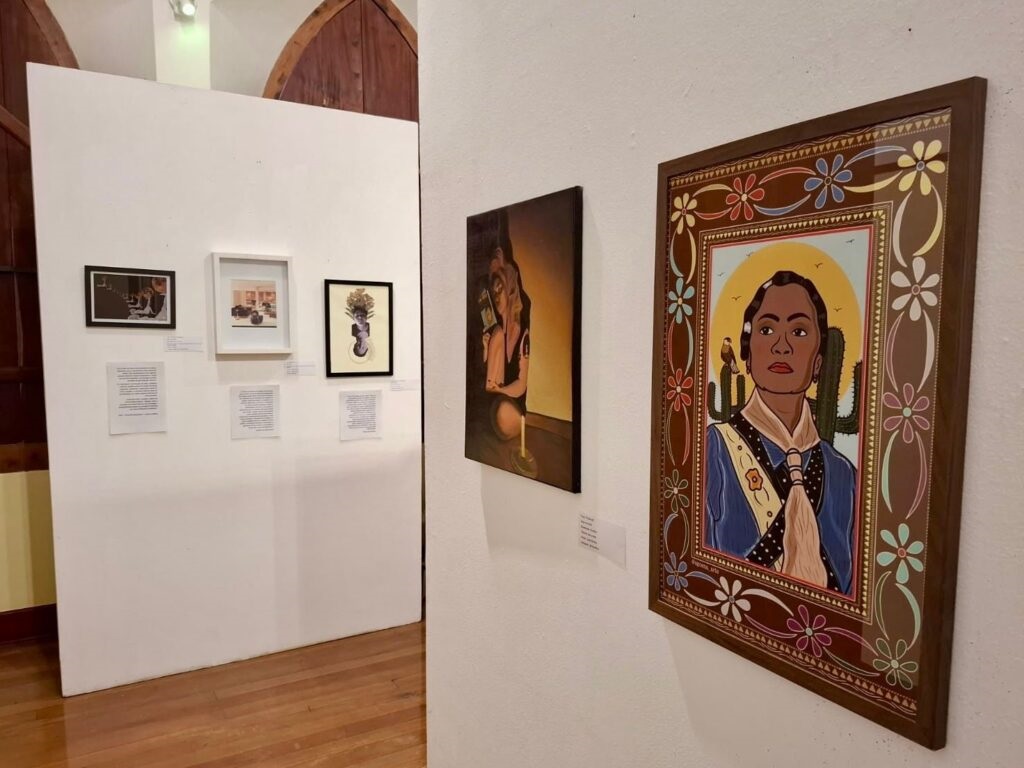 A imagem mostra uma exposição de arte em uma galeria. As obras estão penduradas em paredes brancas, com iluminação suave que destaca as peças. À esquerda, há três quadros: um pequeno em preto e branco no topo, abaixo um quadro colorido, e ao lado direito, uma obra que parece ser uma colagem ou uma pintura com uma silhueta escura de uma cabeça adornada com flores e folhas. Abaixo de cada quadro, há etiquetas brancas, provavelmente com informações sobre as obras. Na parede à direita, há duas pinturas. A mais próxima é um retrato com moldura de madeira ornamentada, mostrando uma pessoa com cabelo escuro, vestindo uma roupa azul e uma faixa, com um halo amarelo ao redor da cabeça e um cacto ao fundo, com um estilo que remete à arte popular, usando cores vibrantes e traços fortes. À esquerda dessa pintura, outra obra está parcialmente visível, mostrando uma figura sob uma luz dourada, com fundo escuro. O chão é de madeira polida, refletindo a luz ambiente. Ao fundo, é possível ver elementos arquitetônicos, como arcos ou janelas com molduras escuras. A composição transmite um ambiente tranquilo e organizado, típico de uma galeria de arte, valorizando as obras expostas.