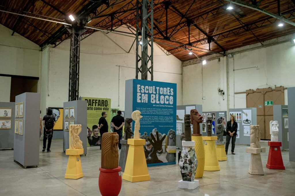 A imagem mostra uma exposição de arte intitulada "ESCULTORES EM BLOCO", do artista Luiz Carlos Fernandes. O espaço expositivo é amplo, com um teto alto de madeira e estrutura metálica aparente, criando um ambiente industrial e moderno. No chão de concreto, estão dispostas várias esculturas estilizadas, que lembram cabeças humanas alongadas e abstratas, cada uma sobre um pedestal colorido — amarelo, vermelho ou branco. No centro, destaca-se uma escultura amarela clara sobre um pedestal amarelo, ao lado de uma escultura marrom escura sobre base amarela com padrão, e outra escultura branca sobre pedestal vermelho. Ao fundo, há painéis cinza com desenhos ou caricaturas emolduradas, e uma grande placa verde com o título "PAFFARO: arte como vida". Algumas pessoas circulam e observam as obras, sugerindo que a exposição está aberta ao público. O texto na parede informa que Luiz Carlos Fernandes é um artista brasileiro nascido em 1959, com vasta experiência como ilustrador, caricaturista, chargista e escultor, tendo trabalhado na imprensa por 40 anos e recebido diversos prêmios nacionais e internacionais. A exposição apresenta um conjunto de suas esculturas, sendo uma mostra representativa de sua carreira artística.