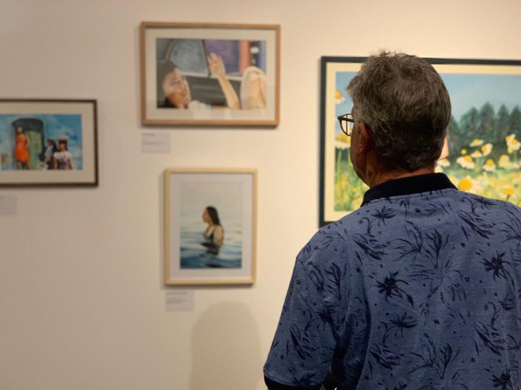 A imagem mostra o interior de uma galeria de arte, onde um homem está observando várias obras expostas na parede. Ele está de costas para a câmera, usando uma camisa azul com estampa de folhas escuras e óculos. Na parede à sua frente, há diversas pinturas emolduradas. As obras variam em estilo e tema: uma mostra duas figuras humanas, outra retrata uma pessoa deitada, possivelmente dentro de um veículo, e uma terceira apresenta uma pessoa na água usando um maiô preto. Há também uma pintura maior que exibe um campo de flores amarelas e brancas, provavelmente margaridas, sob um céu azul. A iluminação da galeria é suave e destaca as cores das pinturas, enquanto a parede branca cria um fundo neutro que realça as obras. O ambiente transmite uma atmosfera de contemplação e apreciação artística.