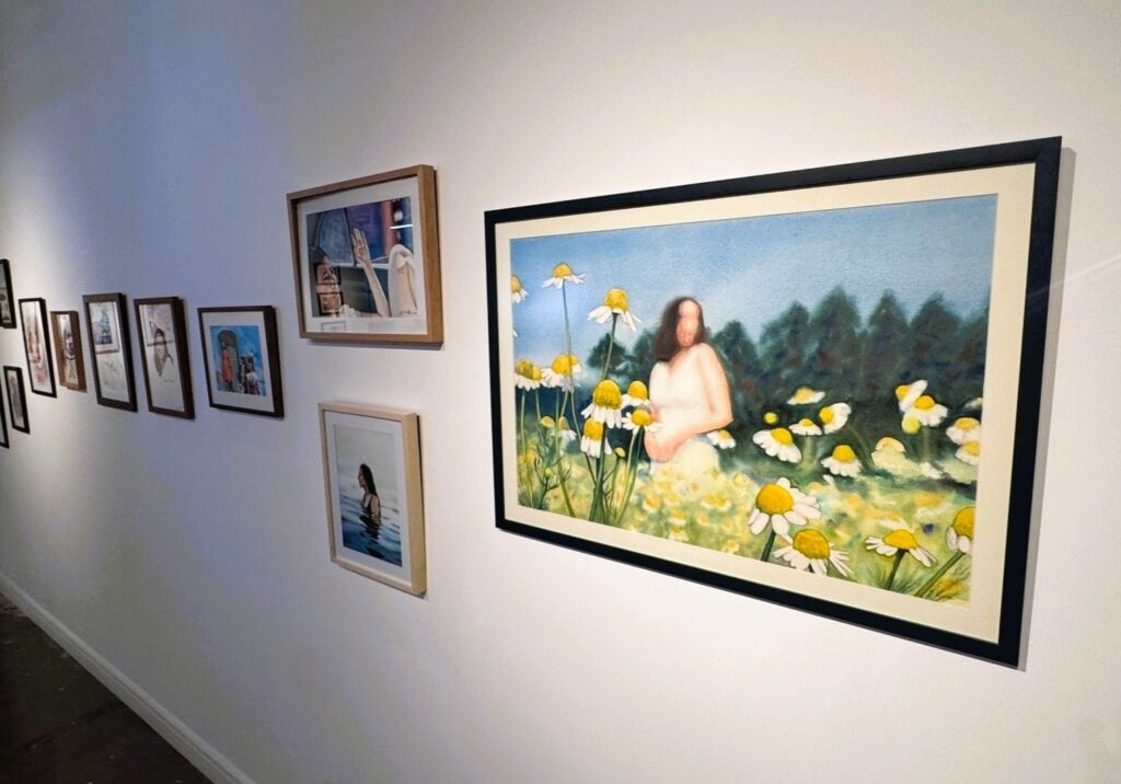 A imagem mostra uma parede branca de uma galeria de arte, onde estão expostas várias obras emolduradas. Em destaque, há uma pintura grande que retrata uma figura humana, possivelmente uma mulher, vestindo um vestido branco e em pé em um campo cheio de margaridas. O fundo da pintura mostra um céu azul e árvores verdes, e a figura está levemente desfocada, conferindo um aspecto etéreo ou impressionista à obra. Ao lado dessa pintura maior, há uma série de quadros menores alinhados na parede, cada um com diferentes estilos e temas, incluindo retratos e cenas figurativas. Um dos quadros menores mostra uma pessoa na água. As molduras variam em tamanho e estilo, criando uma composição organizada, mas diversa. O chão é escuro e contrasta com a parede branca, que serve como um fundo neutro para destacar as obras. A iluminação da galeria realça as texturas e cores das pinturas, e a perspectiva da foto é ligeiramente inclinada para cima, dando profundidade ao espaço exibido.