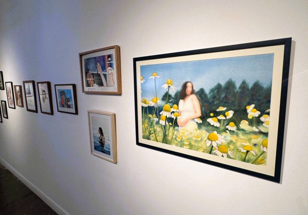 A imagem mostra uma parede de galeria de arte com várias obras emolduradas dispostas em uma linha que se estende da esquerda para a direita, indo do primeiro plano até o fundo. A obra mais destacada está à direita da imagem: uma pintura grande, horizontal, que retrata uma figura humana desfocada, vestindo um vestido claro, em um campo cheio de margaridas. Ao fundo da pintura, há uma floresta verde escura sob um céu azul. A figura da pessoa está sem detalhes nítidos no rosto, transmitindo uma sensação onírica ou etérea. À esquerda dessa grande pintura, há outras obras menores emolduradas. Uma delas mostra uma pessoa na água, talvez nadando ou tomando banho, com uma moldura de madeira clara. Acima dessa, há uma obra com uma moldura de madeira escura, que retrata uma mão levantada diante de uma janela ou abertura, com o fundo desfocado. Mais à esquerda, em direção ao fundo da parede, há uma série de quadros menores, possivelmente ilustrações ou esboços, com diferentes molduras, embora os detalhes sejam difíceis de distinguir por causa da distância e resolução da imagem.