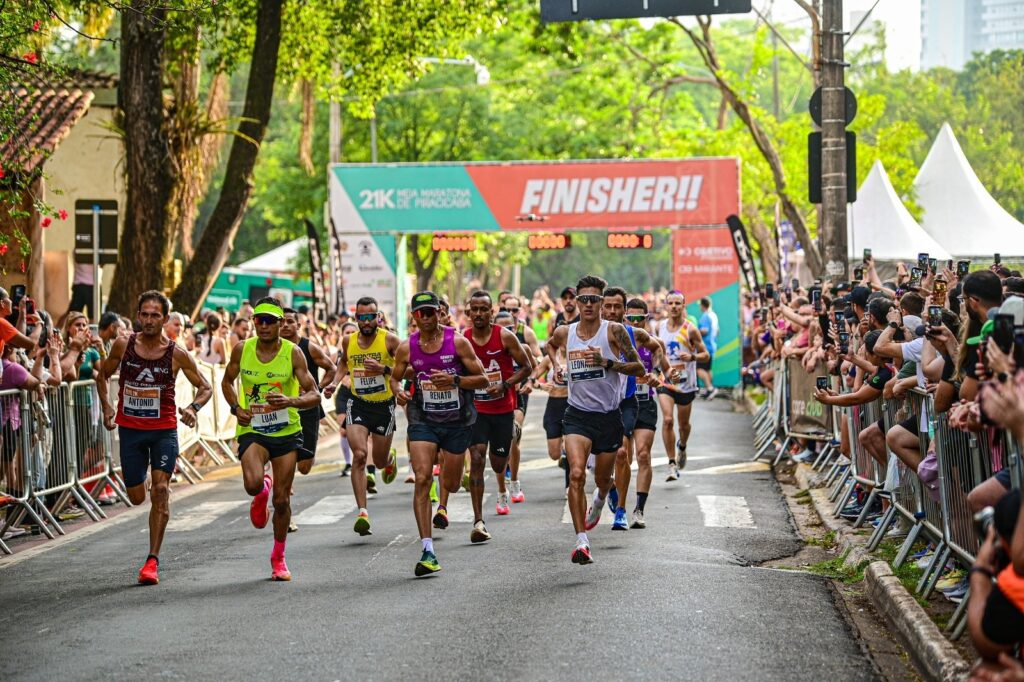 A imagem mostra uma cena vibrante de uma corrida de meia maratona, com vários corredores masculinos em destaque enquanto se aproximam da linha de chegada. Eles estão vestidos com roupas esportivas coloridas, cada um com seu nome visível nas camisetas, como "ANTONIO", "LUAN", "FELIPE", "RENATO" e "LEON". Os corredores exibem expressões de concentração e esforço. Ao fundo, há uma faixa grande que indica o evento: "21K MEIA MARATONA DE PRACAGAMA FINISHER!!", celebrando os participantes que completaram a prova. O local é uma rua arborizada, com árvores verdes formando um cenário natural ao redor. Muitos espectadores estão ao longo do percurso, alguns segurando celulares para registrar o momento. Barricadas metálicas separam o público dos corredores. A atmosfera é de energia e competição, com o sol iluminando a cena, destacando os atletas em movimento e a multidão animada. A imagem transmite o esforço físico dos corredores e a celebração da conquista ao cruzar a linha de chegada.