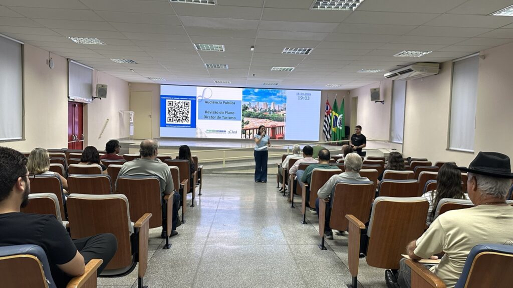 A imagem mostra uma audiência pública realizada em um auditório interno, onde uma mulher está falando em um púlpito no palco, direcionando-se ao público presente. Atrás dela, há uma grande tela que exibe informações sobre o evento, incluindo um código QR, o título "Audiência Pública Revisão do Plano Diretor de Turismo", e a data "15-04-2026" com o horário "19:03". Ao lado da mulher, um homem está em pé, possivelmente auxiliando ou aguardando para falar. O auditório está cheio de pessoas sentadas em cadeiras organizadas em fileiras, todas voltadas para o palco, mostrando interesse na apresentação. As cadeiras têm assentos azuis e o ambiente é iluminado por luzes fluorescentes no teto. Na parede ao lado da tela, estão posicionadas duas bandeiras: a do Brasil e a do estado de São Paulo. O cenário transmite uma atmosfera formal e institucional, típica de eventos públicos e reuniões oficiais. O público é diverso, com homens e mulheres de várias idades, e a disposição do local é organizada para facilitar a visualização e o acompanhamento da apresentação que está sendo feita.