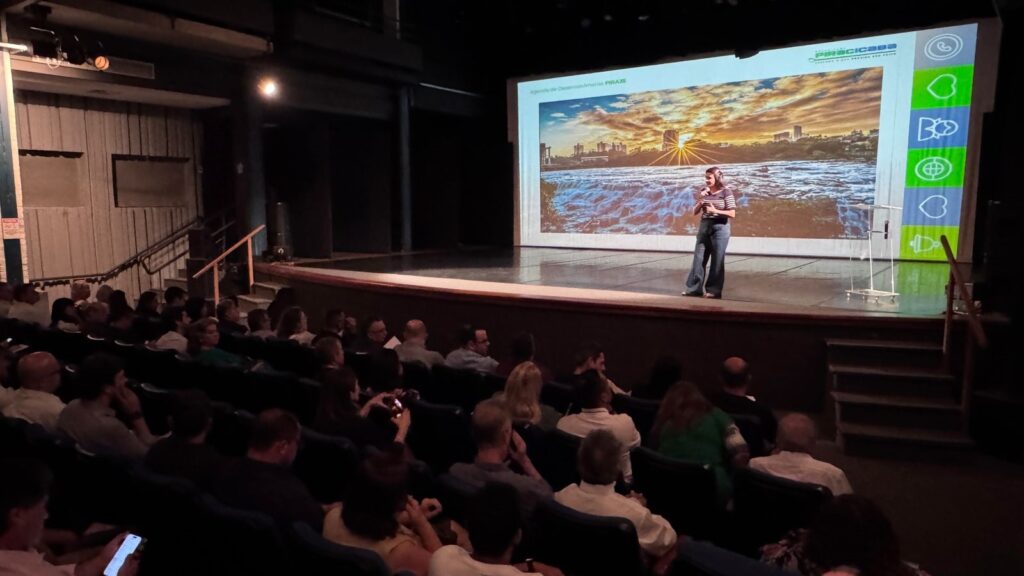 A imagem mostra uma apresentação ocorrendo em um auditório ou teatro. No palco, uma mulher está falando ao microfone, vestindo uma blusa listrada e calça jeans boca de sino. Atrás dela, há uma grande tela de projeção exibindo uma imagem de uma paisagem urbana com um pôr do sol, destacando uma cachoeira e prédios ao fundo. No canto superior direito da tela, está o texto "Agenda de Desenvolvimento PIRACICABA" junto com o logotipo da "PREFEITURA DE PIRACICABA". À direita da tela, há uma faixa vertical com vários ícones, incluindo corações, uma seta circular e um globo. A plateia está sentada em cadeiras alinhadas, assistindo à apresentação. A iluminação do espaço é baixa, com foco de luz na apresentadora e na tela. O ambiente sugere um evento formal ou semi-formal, possivelmente uma reunião ou conferência relacionada ao desenvolvimento urbano ou planejamento da cidade de Piracicaba. A composição da imagem transmite um clima de atenção e interesse por parte do público.