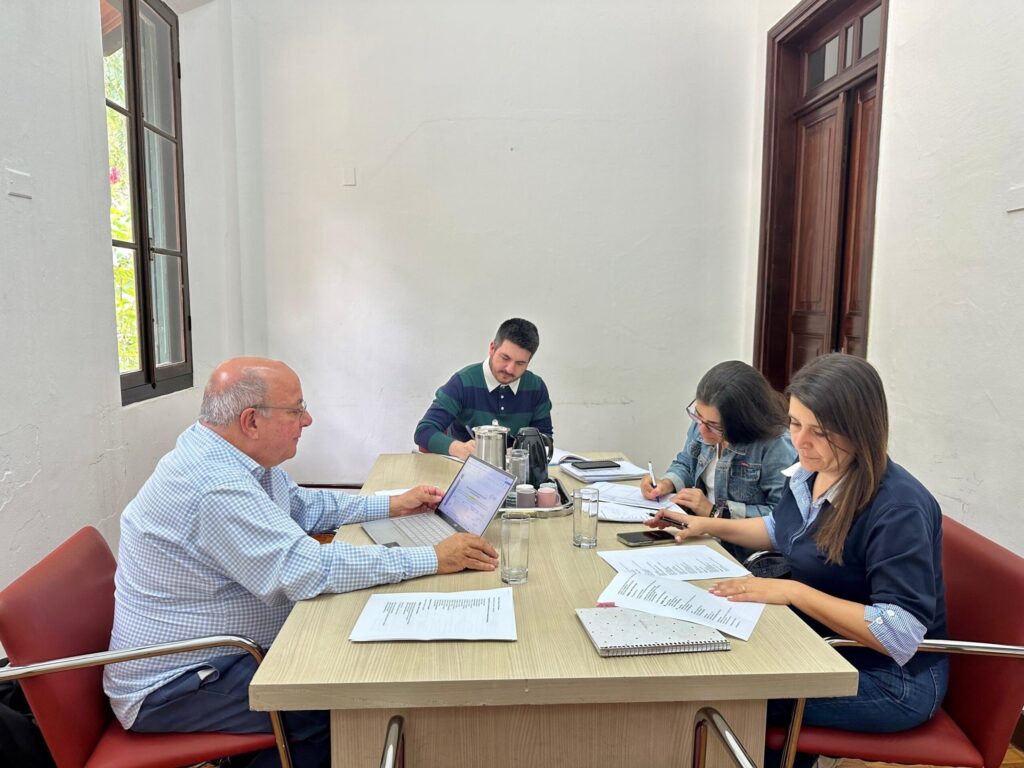 A imagem mostra uma reunião de trabalho em andamento, com quatro pessoas sentadas ao redor de uma mesa retangular de madeira clara. O ambiente é bem iluminado, com luz natural entrando por uma grande janela à esquerda, através da qual é possível ver árvores e vegetação. À esquerda, há um homem mais velho, calvo e usando óculos, vestido com uma camisa xadrez azul clara. Ele está focado em seu laptop aberto à sua frente, aparentemente explicando algo ou apresentando informações para o grupo. Ao fundo, há um homem mais jovem, usando uma camisa listrada verde escura e azul, que está escrevendo em um papel. No centro, uma mulher com óculos e jaqueta jeans está concentrada, também escrevendo ou fazendo anotações. À direita, outra mulher com cabelos longos e escuros, vestindo uma blusa azul escura com gola branca, está lendo ou revisando documentos sobre a mesa. A mesa está organizada com vários papéis, cadernos, copos de água, uma garrafa térmica e um jarro, indicando uma reunião longa ou detalhada. As cadeiras são de couro vermelho, conferindo um toque de sofisticação ao ambiente. O clima geral da imagem transmite colaboração, concentração e profissionalismo, em um espaço que parece ser um escritório ou sala de reuniões com um ambiente acolhedor e iluminado.