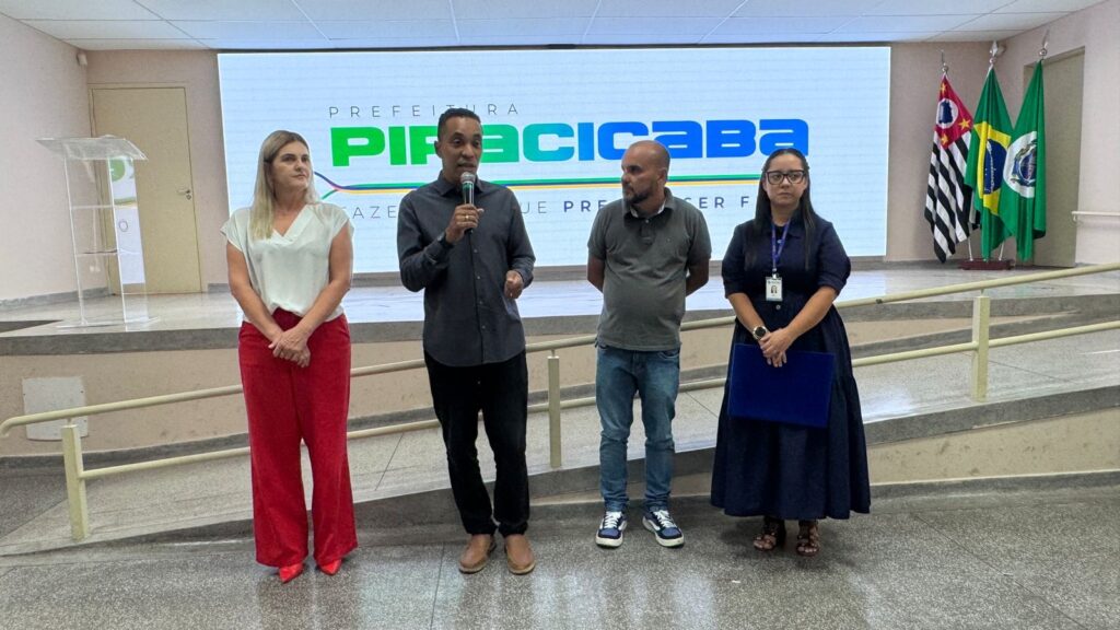 A imagem mostra uma cerimônia oficial realizada em um ambiente interno, possivelmente um auditório ou sala de conferências. Quatro pessoas estão alinhadas em pé, voltadas para a câmera, sobre um palco ou plataforma elevada. Atrás delas, há uma grande tela digital exibindo o logotipo e o nome "PREFEITURA PIRACICABA", acompanhado de elementos gráficos coloridos. Ao lado da tela, estão posicionadas três bandeiras: a bandeira do estado de São Paulo, a bandeira do Brasil e outra bandeira parcialmente visível, provavelmente relacionada ao município. As pessoas na imagem estão vestidas de maneira formal e semi-formal. Da esquerda para a direita: uma mulher com blusa branca e calça vermelha, um homem segurando um microfone e falando, vestido com camisa cinza escura e calça preta, outro homem com camisa polo cinza e jeans, e uma mulher com vestido azul escuro e óculos, segurando uma pasta azul. O ambiente é bem iluminado e a atmosfera parece ser de um evento institucional ou governamental, possivelmente uma apresentação ou anúncio oficial da prefeitura de Piracicaba.