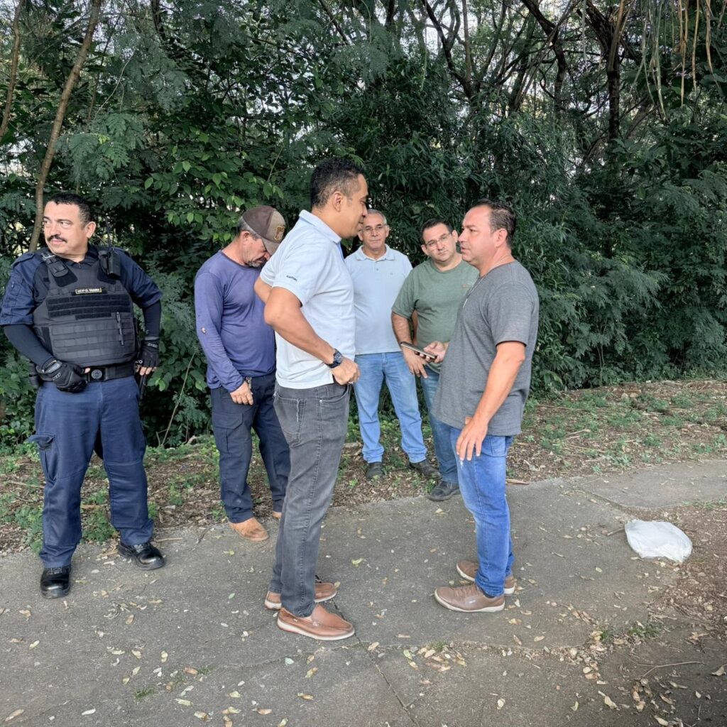 A imagem mostra um grupo de seis homens em um caminho pavimentado ao lado de uma área com muitas árvores e vegetação densa. Um dos homens, à esquerda, está vestido com uniforme policial, incluindo colete tático e luvas pretas, aparentando estar em serviço. Os outros cinco homens estão vestidos de forma casual, com camisetas e calças jeans. Dois deles parecem estar conversando no centro da imagem, enquanto outro segura um celular e observa a conversa. O ambiente parece ser ao ar livre, em um parque ou área verde, com luz natural suave, indicando que pode ser fim de tarde. No chão, há algumas folhas espalhadas e uma sacola plástica branca. A cena transmite uma interação entre os homens, possivelmente relacionada a uma abordagem ou conversa formal, dada a presença do policial.