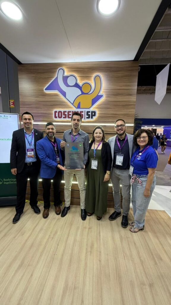 A imagem mostra um grupo de seis pessoas, três homens e três mulheres, posando para uma foto em um ambiente interno, provavelmente em uma feira ou evento corporativo. Eles estão alinhados em frente a um painel de madeira com o logotipo do COSEMS SP (Conselho de Secretarias Municipais de Saúde do Estado de São Paulo) e algumas palavras escritas, como "Cidade Alçacar", "Jardim Botânico", "Cidadania Ativa" e "Nível 1". Uma das pessoas, um homem no centro, está segurando uma placa transparente, que parece ser um prêmio ou reconhecimento. O grupo está vestido de forma casual para negócios, com roupas que variam entre camisas, blazers e vestidos. Ao fundo, é possível ver outros estandes e pessoas, indicando que a foto foi tirada em um evento ou congresso. O ambiente é bem iluminado e o chão é de madeira clara. A atmosfera geral é de celebração e reconhecimento, sugerindo que o grupo recebeu uma premiação ou destaque relacionado ao COSEMS SP.