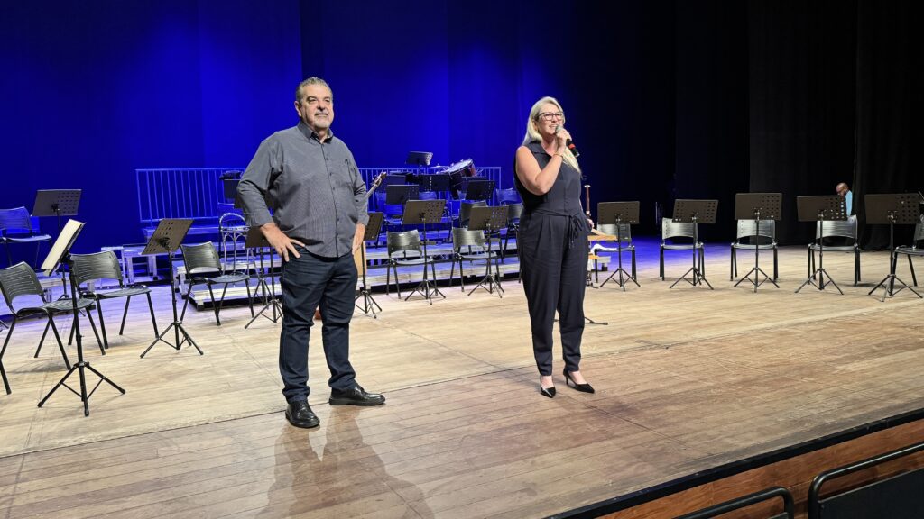 A imagem mostra um palco de madeira com duas pessoas em destaque, um homem e uma mulher. O fundo é dominado por uma cortina azul vibrante, e há várias cadeiras e estantes de partituras organizadas, sugerindo que o local é uma sala de concertos ou um espaço para apresentações musicais. O homem, à esquerda, está de pé com as mãos na cintura, usando uma camisa escura com estampa e calças jeans escuras. Ele tem cabelo grisalho e bigode. A mulher, à direita, veste um macacão escuro, está segurando um microfone e parece estar falando. Ela usa óculos e tem cabelo loiro preso. A iluminação foca nos dois, criando sombras no chão de madeira. A cena transmite a ideia de um evento ou ensaio musical prestes a acontecer ou em andamento.