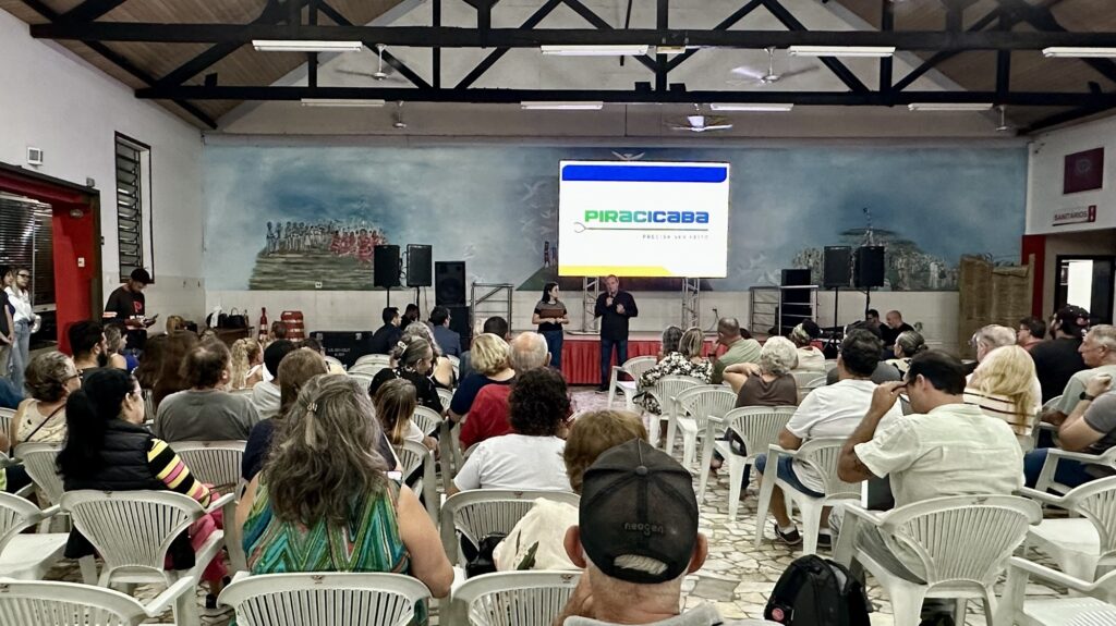 A imagem mostra uma reunião comunitária ou apresentação realizada em um salão amplo com teto alto e estrutura de madeira escura aparente. O ambiente é iluminado por várias lâmpadas industriais penduradas no teto. No centro da imagem, um grupo de pessoas está sentado em cadeiras plásticas brancas, todas voltadas para o palco à frente. No palco, um homem está falando ao microfone, enquanto uma mulher está ao seu lado, aparentemente auxiliando ou aguardando para falar. Ao fundo do palco, há uma grande tela digital exibindo o logotipo "PIRACICABA" acompanhado do slogan "PRECISA SER FEITO". Atrás da tela, na parede, há um mural grande e antigo que retrata pessoas em um cenário natural. O salão tem paredes brancas e piso de cerâmica clara. À direita, uma porta com uma placa indicando "SANITÁRIOS" é visível. O público é diversificado, composto por homens e mulheres de várias idades, atentos à apresentação. O cenário sugere um evento formal de caráter comunitário ou institucional, possivelmente uma reunião pública, palestra ou assembleia.