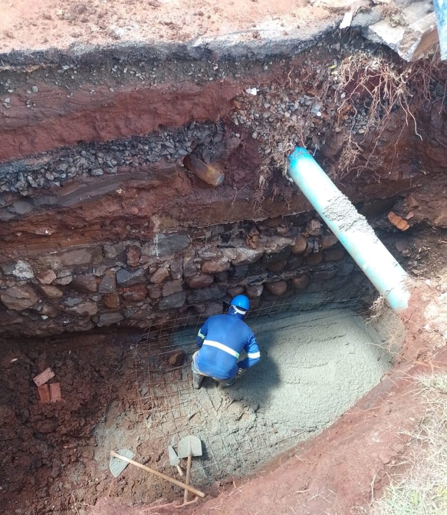 A imagem mostra um trabalhador da construção civil dentro de uma escavação profunda. Ele está agachado, usando um capacete azul e um uniforme azul com faixas refletivas. No chão da escavação, há uma malha de aço (vergalhão) e uma área com concreto fresco sendo despejado por um tubo de PVC azul claro que se estende da direita para o centro da imagem. A parede da escavação revela diferentes camadas de solo, cascalho e uma base de pedras. No primeiro plano, sobre o solo, estão duas pás. A cena é iluminada pela luz natural do dia e parece representar um trabalho de fundação ou instalação de tubulação em um canteiro de obras ao ar livre.
