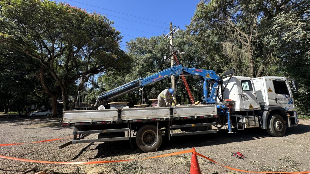 A imagem mostra uma cena de trabalho ao ar livre, em um ambiente que parece ser próximo a uma área arborizada. No centro da imagem, há um caminhão branco equipado com um guindaste azul, identificado pela marca "MOSAL MS 1600S". No caminhão, há grandes bobinas de cabos elétricos, e um trabalhador vestido com uniforme claro e capacete azul está manipulando esses cabos. Ao fundo, é possível ver postes de energia elétrica com fios, indicando que o serviço está relacionado à manutenção ou instalação da rede elétrica. Também há uma área delimitada por fita de segurança laranja no primeiro plano, sugerindo que o local está isolado para o trabalho. À direita, um carro branco está estacionado, e a vegetação verde com algumas flores roxas compõe o cenário. O céu está claro e azul, indicando um dia ensolarado. No caminhão, há inscrições que indicam a empresa "eletrorede cp paulista" e um número de telefone gratuito "0800 0000". O ambiente transmite uma sensação de organização e segurança, com o trabalhador utilizando equipamentos de proteção e o local devidamente sinalizado.