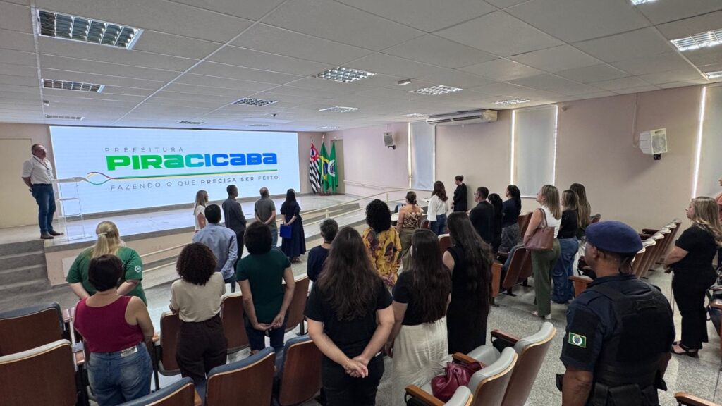 A imagem mostra uma reunião ou evento formal realizado em um auditório ou sala de conferências. Na frente da sala, há uma grande tela de projeção exibindo o texto "PREFEITURA PIRACICABA FAZENDO O QUE PRECISA SER FEITO", junto a um logotipo. Ao lado da tela, estão posicionadas três bandeiras, provavelmente representando o município, o estado e o país. O ambiente está bem iluminado e cheio de pessoas, a maioria delas de pé, voltadas para a tela, indicando que estão prestando atenção a uma apresentação ou discurso. As cadeiras estão organizadas em fileiras, e algumas pessoas estão sentadas enquanto outras permanecem em pé. Em primeiro plano, é possível ver a figura de um indivíduo com uniforme azul escuro e boina azul, que aparenta ser um segurança ou agente de segurança do evento, observando a plateia. A atmosfera parece formal e respeitosa, possivelmente um evento oficial da prefeitura de Piracicaba, com foco em temas relacionados à administração pública ou à comunidade local.