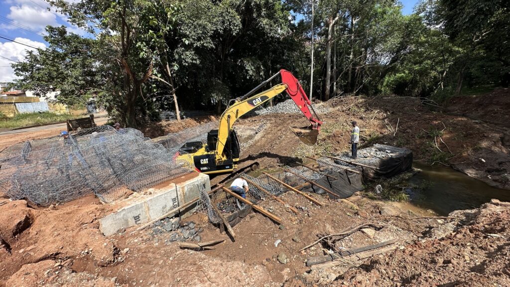 A imagem mostra um canteiro de obras próximo a um pequeno curso d'água, possivelmente um riacho. No centro da cena, há uma escavadeira amarela da marca Caterpillar, modelo 318D L, que está em operação, cavando e movimentando terra com sua caçamba. Dois trabalhadores estão presentes: um deles está próximo à água, aparentemente trabalhando na estrutura ou fundação, vestindo uma camisa branca e calça jeans; o outro está em um monte de terra, observando ou coordenando, usando uma camisa clara e calça escura. A obra parece envolver a construção ou reforço de uma estrutura com gabiões (cestas de arame preenchidas com pedras) e concreto. O solo é de terra vermelha, típico de regiões mais secas ou áridas, e ao fundo há uma vegetação verde densa, com muitas árvores, indicando uma área natural preservada. O céu está parcialmente nublado, e a iluminação sugere que é durante o dia, possivelmente no fim da manhã ou início da tarde, com sombras bem definidas. A palavra "BLO" está visível em uma estrutura de concreto na parte direita da imagem. A perspectiva da foto é levemente elevada, olhando para baixo, capturando bem a atividade e o ambiente ao redor. A cena transmite um momento ativo de construção, com destaque para a interação entre a máquina, os trabalhadores e a natureza.