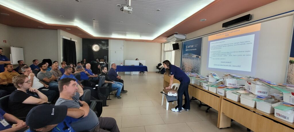 A imagem mostra uma sala de treinamento onde uma mulher, vestindo uniforme azul escuro, está demonstrando técnicas de reanimação cardiopulmonar (RCP) em um manequim colocado sobre uma cadeira. Ela está inclinada sobre o manequim, com as mãos posicionadas no peito, simulando compressões torácicas. Na sala, cerca de 20 a 25 pessoas, principalmente homens, estão sentadas em cadeiras organizadas em fileiras, observando atentamente a demonstração. Eles parecem ser funcionários ou participantes de um curso, muitos usando camisas azuis com o logotipo da SEMAE, organização responsável pelo treinamento. Na parede à frente, há um telão projetando informações sobre parada cardiorrespiratória (PCR), com instruções passo a passo para identificar e agir em casos de PCR, como verificar a respiração, pulsos e chamar ajuda ligando para o número de emergência 192. Também há uma mesa com diversos materiais médicos e caixas, possivelmente para prática ou demonstração. O ambiente é uma sala de conferência ou treinamento, com luz artificial clara, paredes claras, um quadro branco ao lado e cortinas escuras ao fundo. A atmosfera é profissional e focada na segurança e no aprendizado de primeiros socorros. O logotipo da SEMAE aparece visível no telão e em algumas roupas, reforçando a identidade da organização que promove o curso.