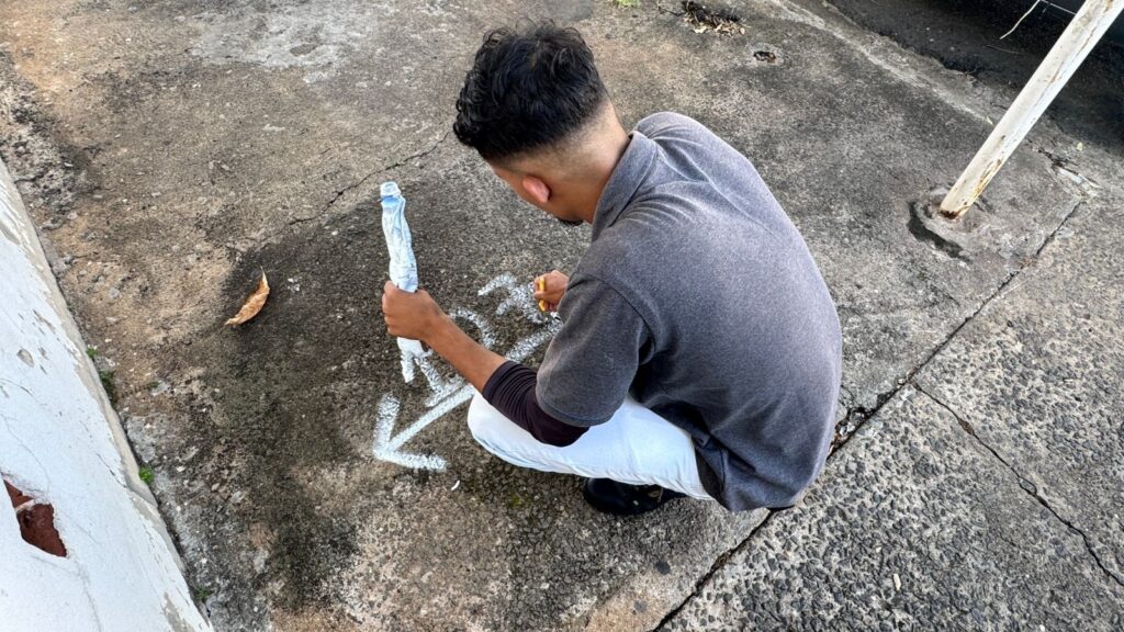 A imagem mostra um homem visto por trás, agachado enquanto desenha no chão de concreto. Ele tem cabelo escuro e encaracolado, veste uma camisa polo cinza com mangas escuras e uma calça branca. Na mão esquerda, segura uma garrafa plástica amassada, e na mão direita, está usando um marcador ou giz amarelo para fazer desenhos brancos no chão. No concreto, é possível ver algumas marcas brancas que parecem setas ou formas abstratas. O ambiente parece ser ao ar livre, com o chão desgastado e algumas folhas secas espalhadas. Há também um poste branco enferrujado à direita do homem. A cena está iluminada pela luz do dia, com sombras visíveis no chão. A imagem transmite a ação de criar algo no espaço urbano, destacando o contato entre o homem e a superfície onde ele está desenhando.