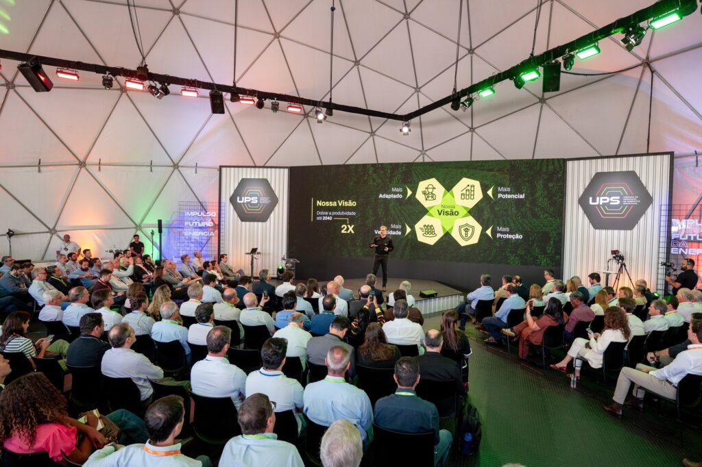 A imagem mostra uma apresentação realizada dentro de uma grande estrutura branca em formato de cúpula geodésica, provavelmente um centro de convenções ou evento. No palco, um homem vestido com camiseta preta está falando para uma plateia sentada, composta por homens e mulheres em trajes casuais e de negócios. Atrás do apresentador, uma grande tela exibe slides da apresentação, com o logotipo da UPS e textos em português. O slide principal apresenta a visão da empresa, destacando pontos como "Mais Adaptado", "Mais Potencial", "Mais Velocidade" e "Mais Proteção". Também há a meta de "Dobrar a produtividade até 2040", com o símbolo "2X". O ambiente é iluminado com luzes coloridas, incluindo verde e vermelho, que estão instaladas na estrutura metálica do teto da cúpula. A plateia está atenta ao palestrante, e o cenário geral transmite uma atmosfera profissional e tecnológica.