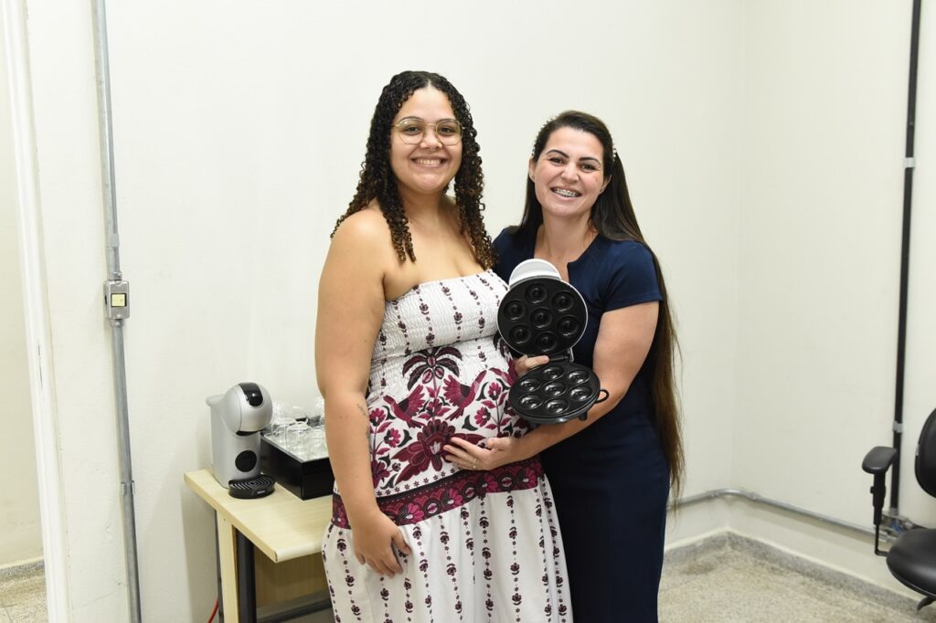 A imagem mostra duas mulheres em um ambiente interno, com uma parede clara ao fundo. A mulher à esquerda está grávida, usa óculos e um vestido branco sem alças com estampa roxa e preta de flores e pássaros. Ela tem cabelos longos e cacheados e está sorrindo para a câmera. A mulher à direita tem cabelos longos e lisos, veste um vestido azul escuro e usa aparelho nos dentes. Ela está sorrindo e segurando uma máquina de fazer donuts aberta, posicionada na barriga da mulher grávida. Ao lado delas, há uma mesa pequena com uma máquina de café prateada e um suporte com copos pequenos. Também é possível ver tubulações aparentes na parede. A cena transmite uma atmosfera alegre e descontraída.