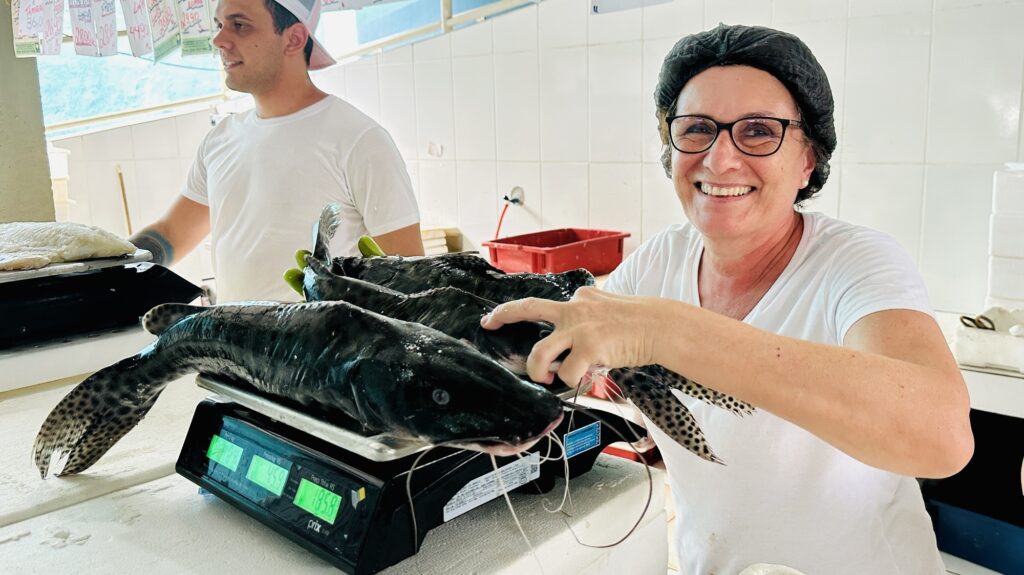 A imagem mostra uma cena em um ambiente interno, provavelmente um mercado ou uma peixaria. Em primeiro plano à direita, há uma mulher sorridente, vestindo uma camiseta branca, uma touca preta e óculos, segurando três grandes bagres (peixes) sobre uma balança digital preta. A balança exibe números em verde, indicando o peso ou preço dos peixes. Ao fundo, à esquerda, um homem com camiseta branca e boné rosa e branco observa algo fora da câmera. O ambiente é limpo, com paredes azulejadas brancas e algumas etiquetas de preços penduradas no teto, onde aparecem valores como "Tambaqui 36,00", "Lombo 26,90" e outros preços em reais. Há também uma caixa plástica vermelha ao fundo. A iluminação é clara, destacando os detalhes dos peixes e o rosto feliz da mulher, transmitindo uma atmosfera de trabalho e comércio de pescado.