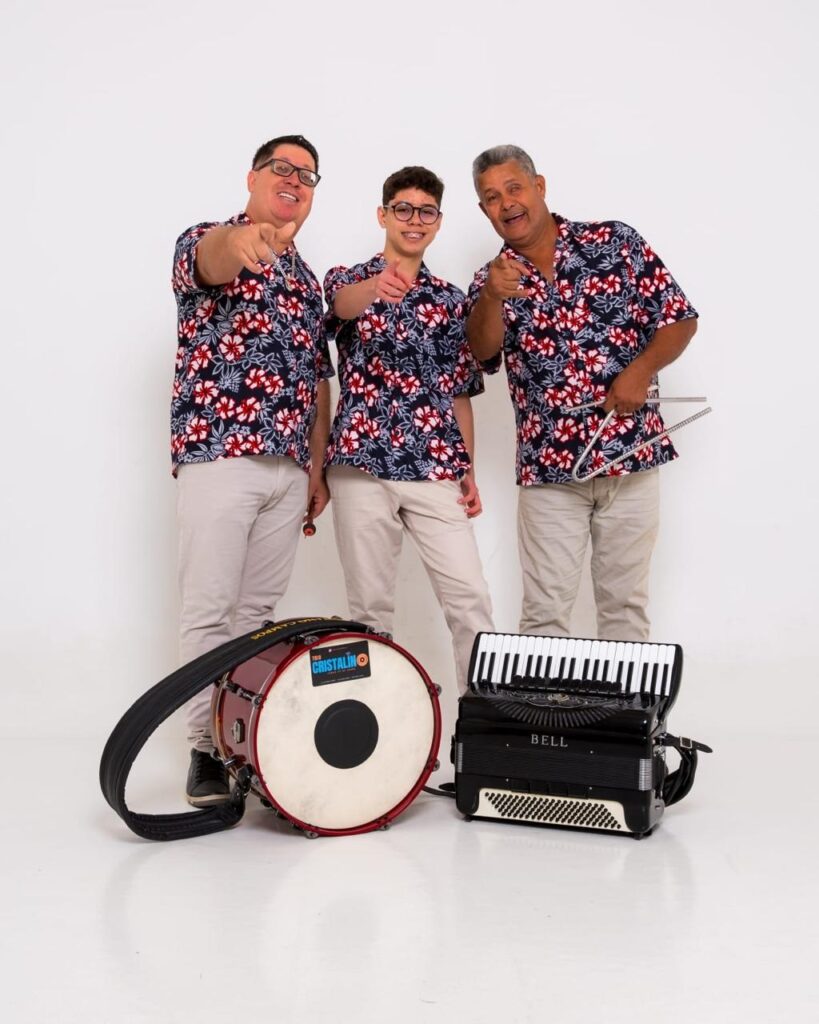 A imagem mostra três homens posando juntos diante de um fundo branco liso. Eles vestem camisas de botão com estampa floral vermelha e branca sobre um fundo azul escuro, combinando com calças claras. Todos estão sorrindo e apontando para a câmera com o dedo indicador, transmitindo uma energia alegre e convidativa. Na frente deles, no chão, há instrumentos musicais: um grande tambor de marcha vermelho com a marca "TRIO CRISTALINO" visível na pele do tambor, um acordeão preto com detalhes brancos e a palavra "BELL" escrita, e um triângulo que o homem da direita está segurando, pronto para ser tocado. A iluminação é clara e uniforme, destacando os detalhes dos instrumentos e das roupas, e o cenário simples ajuda a focar nos músicos e seus instrumentos. A imagem transmite uma sensação de festa e música ao vivo, possivelmente de um grupo musical chamado "Trio Cristalino".