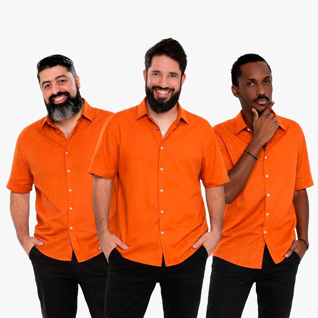 A imagem mostra três homens alinhados lado a lado, todos vestindo camisas laranja de mangas curtas e calças pretas. O homem à esquerda tem uma barba cheia, com fios grisalhos nas bordas, e está sorrindo com as mãos nos bolsos. O homem do meio também possui barba cheia e está sorrindo amplamente, usando um colar prateado. O homem à direita tem uma barba mais curta e está olhando ligeiramente para a esquerda, com a mão tocando o queixo; ele usa uma pulseira no pulso esquerdo. O fundo é branco e a iluminação é clara e uniforme, destacando as cores vibrantes das camisas. A foto é enquadrada do meio do peito para cima, mostrando principalmente os rostos e parte superior dos corpos dos três homens.