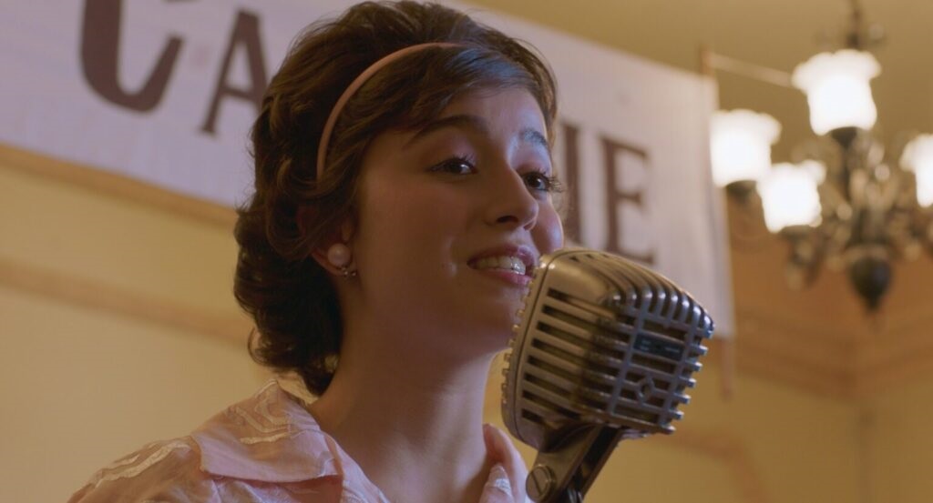 A imagem mostra uma jovem mulher cantando em um microfone vintage. Ela tem cabelos escuros e cacheados, presos por uma faixa rosa clara, que combina com sua blusa delicada, também rosa, com detalhes em renda. Ela usa brincos de pérola e um anel, e sua maquiagem é caprichada, destacando os olhos. O microfone tem um design clássico, cromado, e está posicionado bem próximo à sua boca, indicando que ela está no meio de uma apresentação. Ao fundo, há um banner com letras grandes parcialmente visíveis, formando as letras "C", "A" e "E". Também é possível ver um lustre com várias luzes, desfocado, sugerindo que o ambiente é interno, provavelmente um salão ou palco para apresentações. A iluminação é quente, criando uma atmosfera acolhedora e íntima, e o estilo geral da cena remete às décadas de 1950 ou 1960, tanto pelo visual da mulher quanto pelo tipo de microfone. A composição da imagem foca principalmente na cantora, com o fundo desfocado para dar destaque a ela, transmitindo a sensação de um momento de performance musical.