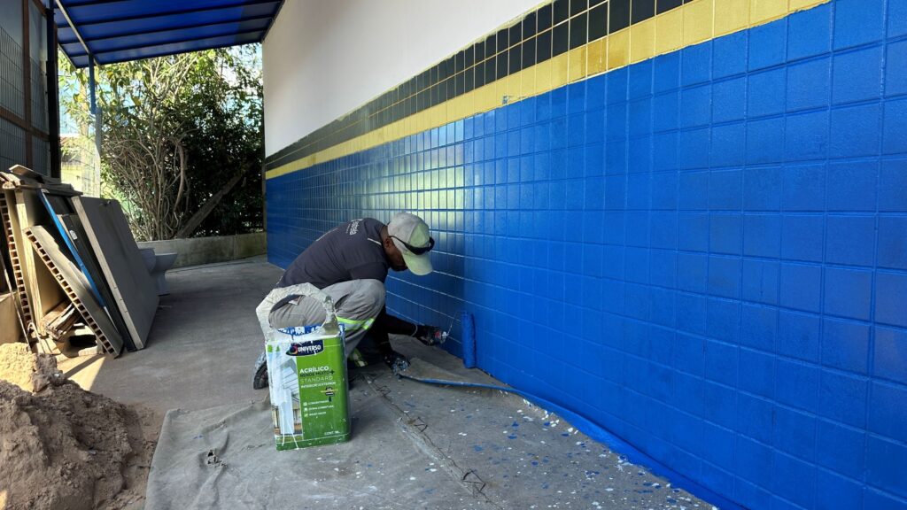 A imagem mostra um trabalhador pintando uma parede de azulejos com tinta azul vibrante. Ele está agachado, usando uma camiseta cinza com a palavra "estrela" nas costas, calças de trabalho com faixas refletivas, luvas pretas, boné claro e óculos escuros apoiados no boné. Ao lado dele, no chão, há um balde grande de tinta verde da marca "Universo", que indica ser tinta acrílica, de alta cobertura, para uso interno e externo, com baixo odor e embalagem de 18 litros. A parede que está sendo pintada é longa e já está parcialmente coberta com a tinta azul, com faixas amarelas e pretas acima do azul, sugerindo que o local pode ser uma instituição pública ou escola, dado o padrão de cores. No fundo, há árvores verdes e uma estrutura com toldo azul, indicando que o local é ao ar livre ou semiaberto. Ao lado esquerdo da imagem, empilhados, há materiais de construção e uma pilha de terra, indicando que o local está em reforma ou manutenção. A luz natural ilumina a cena, criando sombras suaves no chão.