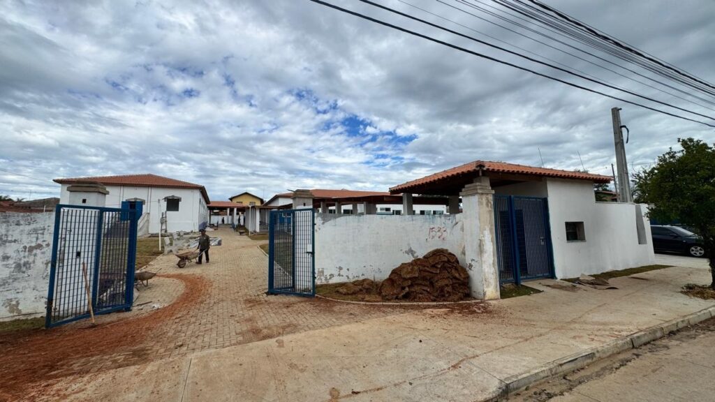A imagem mostra a entrada de um complexo de edifícios, possivelmente uma escola ou clínica, cercado por um muro branco com portões de metal azuis. No pátio interno, um trabalhador está empurrando um carrinho de mão sobre um caminho pavimentado. Os edifícios têm paredes brancas e telhados com telhas laranja. Há uma pilha grande de material marrom, que parece ser terra ou grama, próxima a uma das paredes internas. O céu está parcialmente nublado, com nuvens brancas e cinzas, e a iluminação sugere um dia nublado. No canto direito da imagem, é possível ver parte de um carro escuro estacionado. A cena transmite uma sensação de trabalho em andamento, possivelmente reformas ou manutenção no local.
