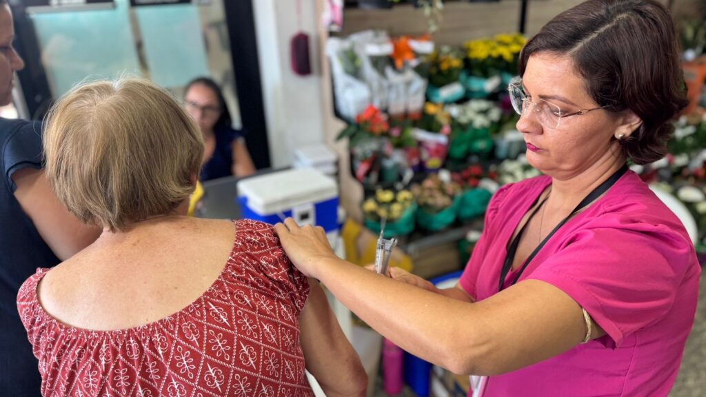 A imagem mostra uma profissional de saúde, vestindo um uniforme rosa vibrante, aplicando uma vacina no braço de uma mulher idosa. A mulher idosa está de costas para a câmera, usando uma blusa vermelha com estampa. A profissional está concentrada na aplicação, segurando a seringa com cuidado. Ao fundo, há uma bancada com um cooler azul e branco, provavelmente usado para armazenar as vacinas, e algumas prateleiras com flores e produtos, indicando que o local pode ser uma farmácia ou clínica de saúde. A iluminação é clara e o foco da imagem está na interação entre as duas mulheres, com o fundo desfocado para destacar o momento da vacinação.