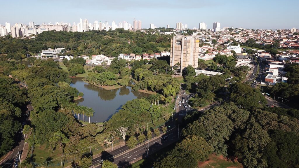 A imagem mostra uma vista aérea de uma área urbana com um grande parque ao centro. No parque, há muitas árvores verdes, incluindo algumas palmeiras, e um lago que reflete o céu azul. Ao redor do parque, há diversas construções residenciais, principalmente casas e prédios baixos. Mais ao fundo, é possível ver prédios maiores, típicos de uma cidade. Há uma estrada que passa próxima ao parque, com alguns carros visíveis. O céu está claro, indicando um dia ensolarado, e a luz do sol cria sombras suaves. A cena transmite uma sensação de equilíbrio entre a natureza do parque e a urbanização ao redor.