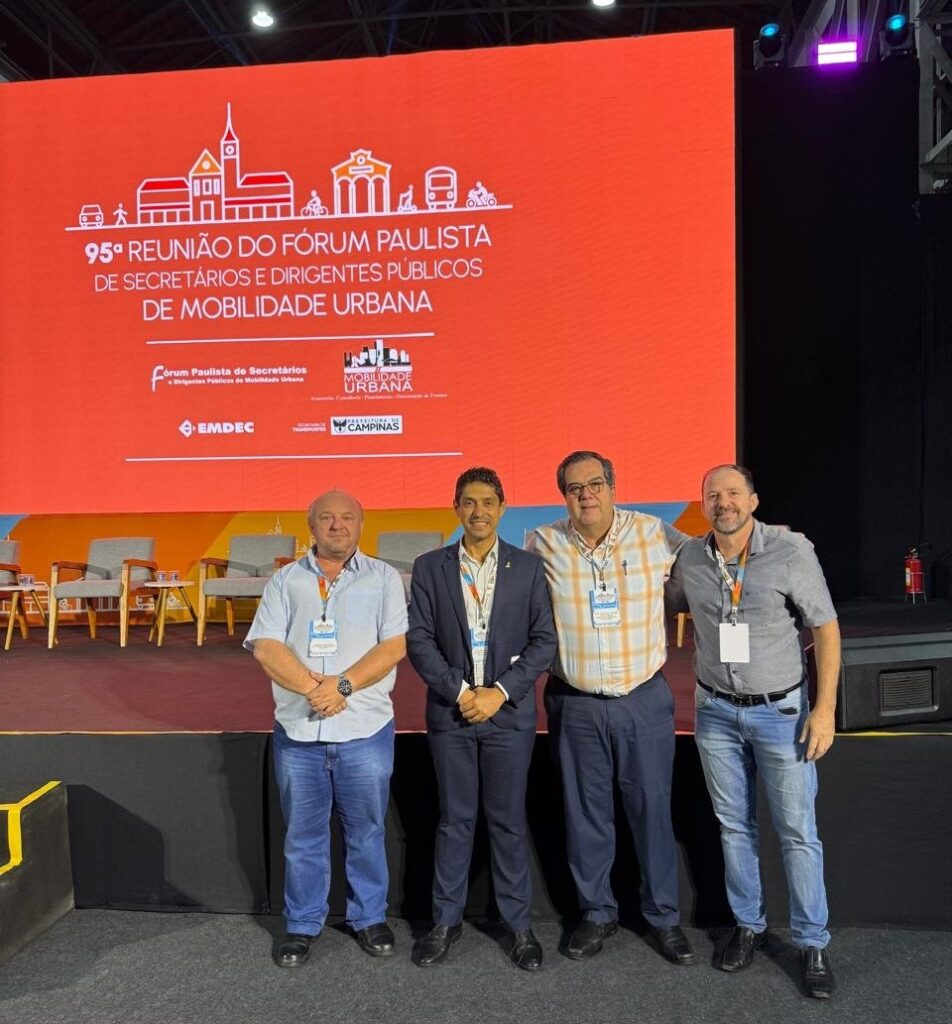 A imagem mostra quatro homens adultos em pé, alinhados, posando para uma foto em um evento formal. Eles estão sobre um palco com um fundo iluminado em laranja, que exibe o título "95º REUNIÃO DO FÓRUM PAULISTA DE SECRETÁRIOS E DIRIGENTES PÚBLICOS DE MOBILIDADE URBANA" junto com logotipos de organizações como EMDEC, Prefeitura de Campinas e Fórum Paulista de Secretários e Dirigentes Públicos de Mobilidade Urbana. Os homens vestem roupas casuais a semi-formais: três usam camisas sociais (uma azul clara, uma xadrez e uma cinza) e um está com um paletó escuro por cima da camisa branca. Todos usam crachás com cordão no pescoço, indicando que são participantes do evento. O cenário sugere que a reunião é relacionada à mobilidade urbana, possivelmente discutindo políticas públicas ou estratégias para transporte nas cidades paulistas. O ambiente é profissional e o foco está na importância do encontro.