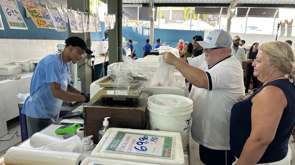 A imagem mostra uma cena movimentada dentro de um mercado de peixes. No centro, um vendedor está atendendo um casal de clientes. O vendedor veste uma camiseta azul, boné preto e um avental plástico transparente, enquanto o homem cliente usa boné branco, camiseta branca e óculos, e a mulher ao lado dele está com uma blusa azul escura. Eles estão em frente a um balcão onde há uma balança digital e vários recipientes, incluindo uma caixa de isopor e um balde branco. No teto, pendurados, há vários cartazes com preços de diferentes tipos de peixe, como "Dourada em postas 69,90 kg", "Posta de tilápia 7,50 kg" e "Bacalhau desfiado 69,90 kg". O ambiente é bem iluminado, com luz natural entrando, e ao fundo é possível ver outras pessoas circulando, o que reforça a ideia de um mercado público movimentado. A imagem tem uma perspectiva realista e foco nítido nos personagens principais.