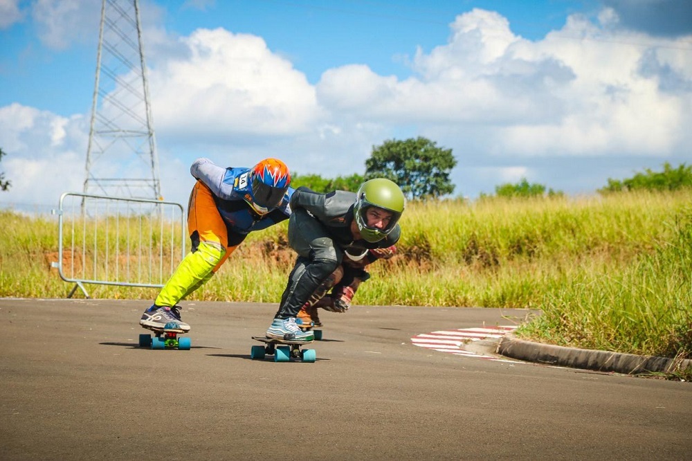 A imagem mostra dois praticantes de longboard descendo uma pista asfaltada em alta velocidade, inclinados para fazer uma curva. Eles usam capacetes e roupas de proteção, evidenciando a natureza esportiva e de risco da atividade. O cenário ao redor é composto por colinas verdes e árvores, sob um céu azul com algumas nuvens. À direita da pista há uma faixa pintada de vermelho e branco, típica de circuitos de corrida, e uma barreira metálica separa a pista da área verde. Ao fundo, uma torre de transmissão elétrica se destaca contra o céu, misturando elementos naturais e urbanos. A perspectiva baixa da foto ressalta a sensação de velocidade e a intensidade da competição entre os dois longboarders.