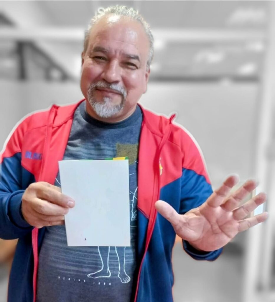 A imagem mostra um homem de meia-idade com cabelos grisalhos e cavanhaque, sorrindo e olhando diretamente para a câmera. Ele está vestindo uma jaqueta azul com detalhes vermelhos por cima de uma camiseta escura com um desenho gráfico que parece ser uma silhueta de pernas e pés. Na mão esquerda, ele segura uma folha de papel branca em branco, enquanto com a mão direita faz um gesto de palma aberta voltada para a câmera. O fundo está desfocado, sugerindo um ambiente interno, possivelmente um escritório ou ginásio, com paredes de cor neutra e formas indistintas. A iluminação é clara e uniforme, destacando o rosto do homem e os objetos que ele segura. A composição é um plano médio, focando no homem e em seu gesto. A imagem transmite uma sensação amigável e acolhedora.