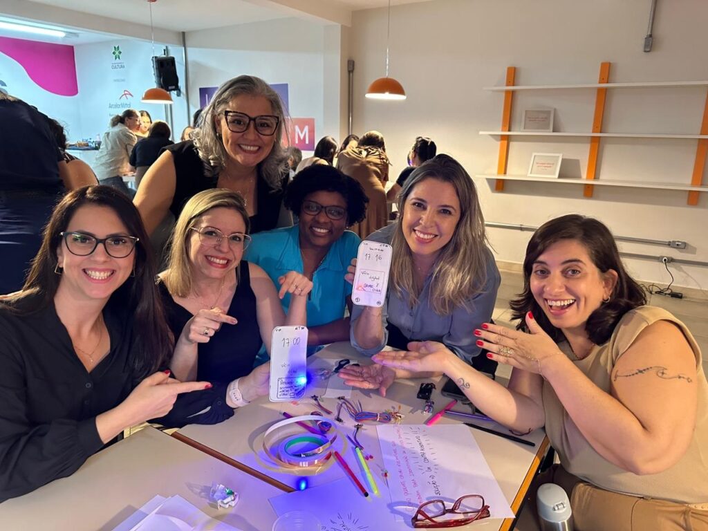 A imagem mostra um grupo de seis mulheres reunidas em torno de uma mesa, participando de uma atividade ou workshop. Elas estão sorrindo e parecem estar animadas e engajadas. Sobre a mesa, há diversos objetos, como papéis, canetas, copos e componentes eletrônicos com fios coloridos, sugerindo que estão trabalhando em um projeto prático ou atividade criativa. Duas das mulheres estão segurando smartphones com a tela visível, mostrando horários e algumas anotações manuscritas. O ambiente parece ser um espaço moderno e bem iluminado, com paredes brancas e prateleiras laranjas ao fundo. Também é possível ver algumas marcas e textos nas paredes, incluindo "CULTURA" e "ArcelorMittal", indicando que o local pode estar relacionado a essa empresa ou a um programa cultural ou educacional promovido por ela. No geral, a imagem transmite uma atmosfera de colaboração, aprendizado e descontração.