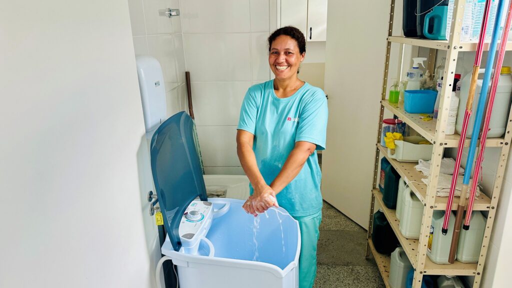 A imagem mostra uma mulher sorridente lavando as mãos em uma pequena máquina de lavar portátil branca com tampa azul. Ela está vestindo uma camiseta azul clara e calças combinando, e parece estar em um ambiente interno, possivelmente uma área de serviço ou lavanderia, com paredes brancas e azulejos. Ao lado da máquina, há uma prateleira metálica cheia de produtos de limpeza e utensílios, como garrafas e esfregões. A iluminação é clara e brilhante, transmitindo uma sensação de limpeza e organização. A máquina de lavar tem a indicação "10KG" em seu painel frontal. A mulher parece estar satisfeita e confortável no ambiente.