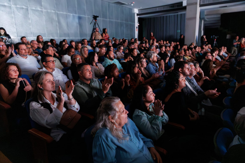 A imagem mostra uma grande plateia sentada em um auditório pouco iluminado, voltada para um palco ou ponto focal que não aparece na foto. A perspectiva é de dentro da plateia, olhando ligeiramente para cima e através das fileiras de pessoas. A iluminação é dramática, com holofotes que iluminam partes da plateia, criando um forte contraste entre luz e sombra. As cores predominantes são tons frios, especialmente azuis das cadeiras, combinados com tons escuros das roupas e do ambiente, além de alguns tons mais quentes provenientes das peles e roupas das pessoas. Muitas pessoas estão aplaudindo, com as mãos levantadas em movimento, e suas expressões são variadas, mas em geral demonstram atenção e envolvimento, olhando para frente ou para cima com olhares focados. A plateia é diversa em termos de idade e etnia. Ao fundo, é possível ver uma câmera em um tripé, indicando que o evento está sendo gravado ou transmitido. O ambiente é moderno, com paredes texturizadas e teto alto. A foto tem boa qualidade, com o fundo desfocado para destacar melhor as pessoas na frente e no meio da plateia. A composição transmite bem a atmosfera de um evento ao vivo e o engajamento coletivo do público.