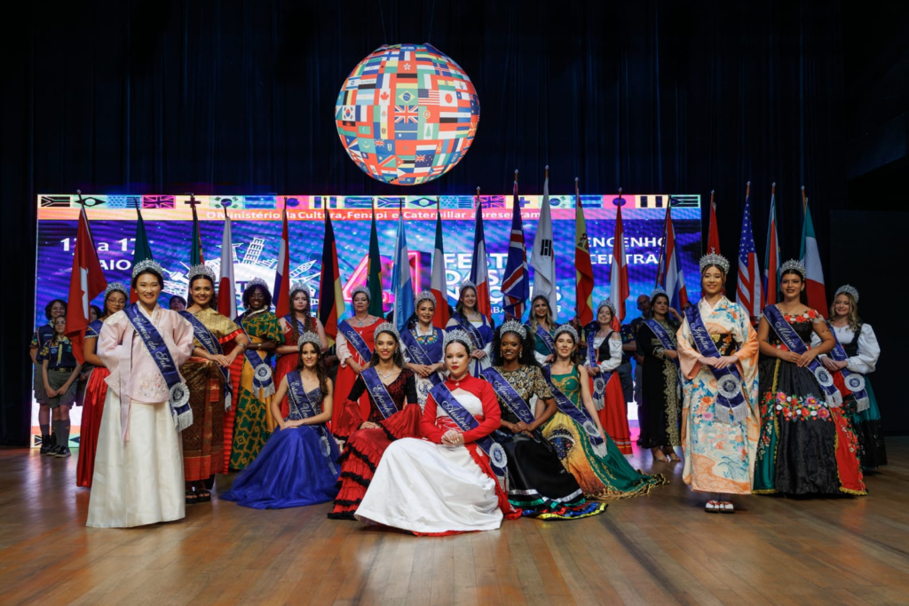 A imagem mostra um grupo de mulheres vestidas com trajes tradicionais de diversas culturas, posicionadas em um palco. Muitas delas usam faixas com nomes de países, como "Coreia do Sul", "Estados Unidos", "África do Sul", "Holanda", "Áustria", "Espanha" e "México", indicando que representam essas nações. Algumas mulheres também usam coroas, sugerindo que se trata de um concurso de beleza ou evento cultural. Ao fundo, há uma grande tela com gráficos coloridos e bandeiras de vários países, além de bandeiras reais em suportes. A iluminação do palco é clara, destacando as participantes e seus trajes típicos, que apresentam uma grande variedade de estilos e cores. A atmosfera geral é festiva e formal, possivelmente um evento internacional que celebra a diversidade cultural.