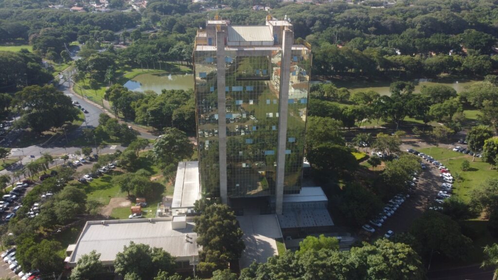 A imagem mostra uma vista aérea de um edifício alto e moderno, com uma fachada de vidro espelhado que reflete a paisagem ao redor, incluindo árvores e o céu. O prédio tem formato retangular e colunas verticais aparentes de concreto nas laterais. Está localizado em uma área verde, com muitas árvores e um pequeno lago ou espelho d’água atrás dele. Em volta do edifício, há estacionamentos com vários carros e vias pavimentadas. A foto foi tirada em um dia claro, com boa iluminação natural, e a perspectiva é de cima para baixo, provavelmente capturada por um drone ou de um ponto elevado. O ambiente ao redor é predominantemente arborizado, transmitindo uma sensação de integração entre a construção e a natureza.